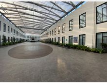 Weitläufiger, moderner Innenhof mit weiß gestrichenen Wänden und großen, rechteckigen Fenstern auf beiden Seiten. Der Raum ist mit einem gläsernen Dach überdacht, bietet dekorative Pflanzen entlang der Seiten und enthält eine runde Holzfläche in der Mitte des Steinbodens.