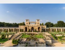 Historisches Gebäude mit symmetrischer Architektur und zentralem Torbogen, flankiert von langen Galerien mit Säulen und Statuen. Im Vordergrund befindet sich ein symmetrisch gestalteter Garten mit Brunnen, Terrassen und Grünflächen, umgeben von Bäumen.