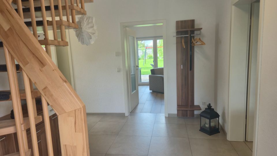 Heller Eingangsflur eines Wohnhauses mit großer Fliesenfläche, weiß gestrichenen Wänden und einer Holztreppe mit Geländer links. Rechts stehen eine schmale Garderobe und eine Laterne am Boden; durch eine offene Tür blickt man in einen Wohnbereich und weiter in den Garten.