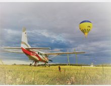 Kleinflugzeug auf einer Wiese mit rot-weißer Lackierung und Kennzeichen "D-FUMP" im Vordergrund; im Hintergrund ein gelb-blauer Heißluftballon am Himmel sowie eine grüne Landschaft unter bewölktem Himmel.