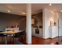 Offener Wohnraum mit integrierter Küche und Essbereich. Dunkler Holzboden, graue Küchenwand mit Schränken, Esstisch mit vier Stühlen und Dekorationen wie Bilder, Pflanzen und Statuen auf weißen Möbeln.