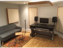 Ein Aufnahmestudio mit schlichter Einrichtung: ein schwarzer Schreibtisch mit Computer, Tastatur und zwei Lautsprechern, ein graues Sofa, ein kleiner Holztisch, ein Mikrofonständer und schallabsorbierende Wand- und Deckenelemente. Der Raum hat helle Holzböden und eine orientalische Teppichdekoration.