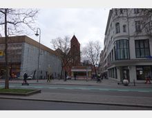 Straßenszene in einer Stadt mit Blick auf ein modernes Gebäude links, ein weißes historisches Gebäude rechts und eine rote Backsteinkirche mit Turm im Hintergrund. Im Vordergrund ein Imbissstand, Fußgänger, ein Elektrorollerfahrer und ein Fahrradweg.
