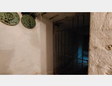 Ein unterirdischer Raum mit groben, weißen Wänden, Metallgittertür und grünen, runden Verschlüssen an der Wand. Im Hintergrund ist ein dunkler Durchgang mit schwachem bläulichem Licht sichtbar.