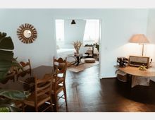 Wohnzimmer und Schlafzimmer in einem offenen, hellen Wohnraum mit Holzboden. Im Vordergrund ein Esstisch mit Holzstühlen, an der rechten Wand ein Beistelltisch mit Lampe und Radio, im Hintergrund ein Schlafzimmer mit einem Bett, rundem Couchtisch und Dekorelementen.