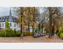 Straßenszene mit herbstlicher Atmosphäre, gesäumt von Bäumen mit gelbem Laub. Im Hintergrund befindet sich ein weißes Gebäude mit spitzem Turmdach und Sprossenfenstern, daneben sind parkende Autos entlang der gepflasterten Straße sichtbar.
