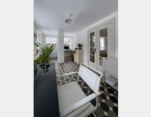 Heller, schmaler Innenraum mit gemustertem Fliesenboden in Grau- und Schwarztönen, weißer Decke und einem zentralen, weiß verkleideten Pfeiler. Am Ende stehen Schreibtisch und Schrank vor großen Fenstern; rechts sind doppelte Glastüren, links eine Fensterbank mit Pflanzen.