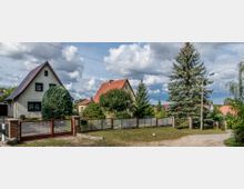 Ein Wohngebiet mit Einfamilienhäusern, darunter ein weißes Haus mit spitzem Dach und ein weiteres mit rotem Ziegeldach. Im Vordergrund ein Garten mit Bäumen und Sträuchern, eine Einfahrt mit einem Zaun und Tor, sowie eine große Tanne.