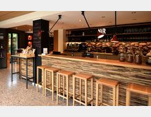 Moderner Café- und Barbereich mit langem Tresen in Holzoptik und hohen Barhockern davor. Im Hintergrund sind Regale mit Gläsern, eine Kaffee-Station, eine mehrfarbige Fliesenrückwand sowie Pendelleuchten und eine Tafelwand zu sehen.