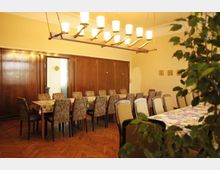 Ein großer, gemütlich eingerichteter Speiseraum mit langen Tischen, die mit farbenfrohen Tischdecken bedeckt sind, und passenden gepolsterten Stühlen. Im Hintergrund befinden sich holzvertäfelte Wände, eine geschwungene Hängelampe mit mehreren Leuchten und ein Parkettboden.