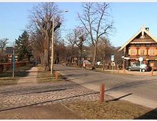 Straßenszene in einem ländlichen Gebiet mit einem Gehweg aus Kopfsteinpflaster, kahlen Bäumen und einem traditionellen Haus im alpinen Stil mit Holzelementen auf der rechten Seite. Im Hintergrund sind mehrere kleinere Gebäude und geparkte Autos sichtbar.