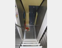 Eine schmale, moderne Treppe aus weißem Metall führt hinunter in einen Flur mit grauem Boden. An den Wänden hängen bunte Bilder, und die Wandverkleidung besteht aus Metall und Glasoberflächen.