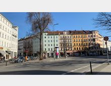 Städtische Straßenszene mit mehrstöckigen Altbauhäusern in Pastellfarben und orangenen Fassaden, Geschäften im Erdgeschoss und geparkten Fahrrädern vor den Gebäuden. Im Vordergrund eine breite Straße mit Verkehrsschildern und Ampeln, sowie ein kahler Baum.