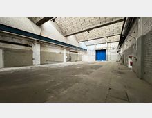 Große leere Lagerhalle mit hohen Decken aus Beton und Wänden aus unverputztem Mauerwerk. Im Hintergrund befinden sich blaue, doppel-flügelige Tore sowie verschiedene Rohrleitungen und Wandbefestigungen.