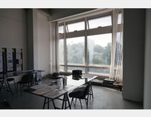 Heller Arbeitsraum mit großen Fenstern, die Tageslicht hereinlassen und einen Blick ins Grüne bieten. Im Raum stehen Tische mit Unterlagen und Büroartikeln sowie einzelne Stühle, und an der Wand ist eine Pinnwand mit Plänen angebracht.