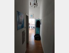 Langer Flur mit hellem Holzfußboden und weiß gestrichenen Wänden, dekoriert mit Postern und gerahmten Bildern. Am Ende des Flurs führt eine Tür in einen anderen Raum mit Dachfenster; im Flur hängen Kleidung und steht eine schwarze Tasche.