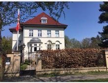Weiße Villa mit rotem Satteldach, symmetrischer Fassade und verzierten Fensterrahmen, umgeben von einer Mauer mit metallischem Gartentor und einer Hecke. Im Vordergrund befindet sich eine Straße, im Hintergrund sind Bäume und ein klarer, blauer Himmel zu sehen.