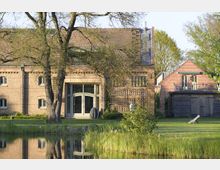Backstein-Landhaus mit großen Fenstern und Holztüren, umgeben von einem gepflegten Garten mit Bäumen, Rasen und einem Teich im Vordergrund. Rechts befindet sich ein Nebengebäude aus Holz und Backstein.