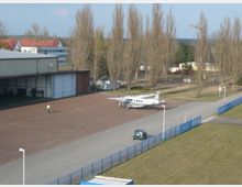 Kleiner Flugplatz mit einer geparkten Propellermaschine auf einer asphaltierten Fläche, daneben ein Hangar mit offenen Toren. Im Hintergrund stehen vereinzelte Bäume, Gebäude mit roten Dächern und ein Zaun, der den Bereich umgibt.
