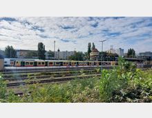 Bahnhofsumgebung mit S-Bahn-Zug auf einem Gleis, der in gelb-rotem Design gestaltet ist. Im Hintergrund sind Wohnhäuser, ein Fachwerkgebäude und Bäume zu sehen, im Vordergrund Büsche und weitere Gleise.