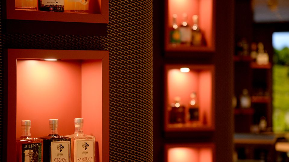 Innenansicht einer Bar mit in die Wand eingelassenen, warm beleuchteten Regalnischen, in denen mehrere Spirituosenflaschen ausgestellt sind. Zwischen den Nischen befindet sich eine dunkle Wandverkleidung mit metallischem Gittermuster, weitere Regale sind unscharf im Hintergrund zu sehen.