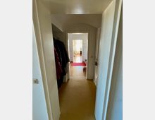 Schlanker Flur in einem Wohnbereich mit beigen Teppichboden und weißen Wänden, links eine Garderobe mit Mänteln, am Ende des Flurs ein hell beleuchteter Raum mit Holzfußboden und rotem Teppich.