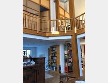 Wohnraum mit hoher Decke und Holzelementen, darunter ein Balkon mit Holzgeländer im oberen Bereich. Im Erdgeschoss sind Bücherregale, ein antikes Holzmöbelstück und ein Esstisch aus Holz sichtbar; helle Wände und große Fenster sorgen für viel Tageslicht.