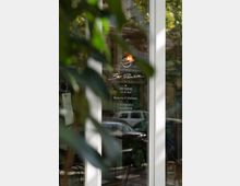 Die Glastür eines Cafés oder Bistros mit der Aufschrift "Bar Panisse" sowie Adresse und Kontaktdaten in Berlin. Im Hintergrund sind Bäume, Blattwerk und verschwommene Fahrzeuge zu sehen.