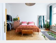 Schlafzimmer mit Parkettboden, in dessen Mittelpunkt ein Doppelbett mit orangefarbener Decke und rosa Kissen steht. Links steht eine Kommode mit Pflanzen und ein Kleiderständer, rechts ein großer Spiegel, ein Teppich und ein Fenster mit türkisfarbenen Vorhängen.