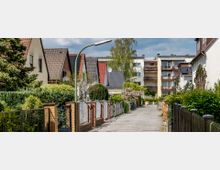 Ruhige Wohnstraße mit Einfamilienhäusern und Mehrfamilienhäusern im Hintergrund, gesäumt von Zäunen, Hecken und blühenden Bäumen. Die Straße ist gepflastert und von modernen Straßenlaternen beleuchtet.