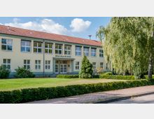 Schulgebäude der Oberschule Premnitz in einem zweigeschossigen, gelb gestrichenen Bau mit rotem Satteldach, großen Fenstern und einem zentralen Eingangsbereich. Vor dem Gebäude befinden sich eine gepflegte Rasenfläche, Büsche, Bäume und ein gepflasterter Weg.