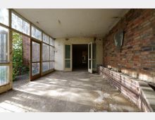 Verlassener Raum mit großen, bodentiefen Fenstern auf der linken Seite, einer Ziegelwand rechts und einer offenen Doppeltür, die in einen dunkleren Innenbereich führt. Der Boden ist aus Beton und zeigt Abnutzungsspuren, während durch die Fenster Tageslicht einfällt.