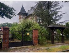 Historische Villa mit Fachwerk-Elementen, spitzem Turmdach und großen Fenstern, umgeben von dichter Vegetation und hohen Bäumen. Im Vordergrund ein eiserner Gartentor mit roten Backsteinpfeilern und einem kleinen Zugang mit Holzüberdachung.