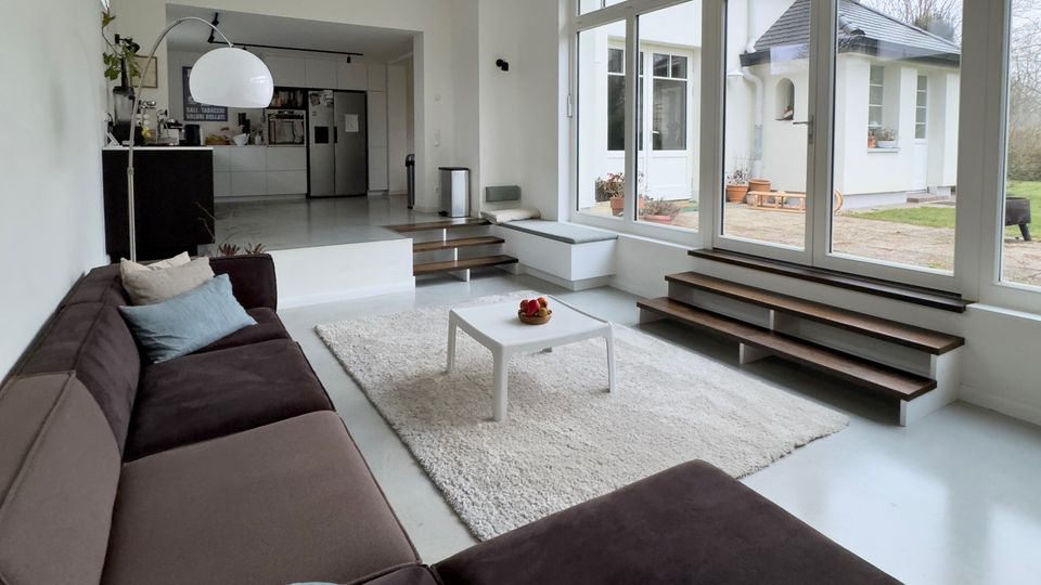 Helles, modernes Wohnzimmer mit großem braunem Ecksofa, weißem Teppich und kleinem Couchtisch, das offen in eine Küche übergeht. Bodentiefe Fensterfront mit seitlichen Stufen führt zur Terrasse und bietet Blick in den Garten und auf ein angrenzendes Haus.