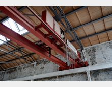 Industriehalle mit offener Holzdachkonstruktion und sichtbaren Stahlträgern. Im Vordergrund ein roter Laufkran an einer Stahlträgerstruktur; die Wände bestehen aus unverputztem, teilweise abblätterndem Mauerwerk.