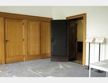 Innenraum mit Holzwandverkleidung und drei geschlossenen Türen aus Holz. Rechts befindet sich eine geöffnete Tür mit dunklem Innenrahmen, daneben stehen zwei beschriftete Schilder auf Metallständern. Der Boden ist mit grauem Teppich belegt, auf dem Glassplitter oder zerbrochene Scheiben sichtbar sind.