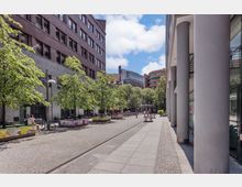 Städtischer Platz mit gepflastertem Boden, flankiert von modernen und klassischen Gebäuden mit Ziegel- und Glasfassaden. Der Platz ist mit Bäumen und dekorativen, bunt bemalten Pflanzkästen gestaltet, mit wenigen Sitzgelegenheiten und Menschen im Hintergrund.