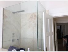 Eine moderne, verglaste Dusche mit beigefarbenen Marmorelementen in einem Badezimmer. Im Hintergrund ist eine offene Tür zu einem Flur mit Holzfußboden sichtbar.