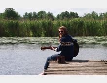 Ein Mann sitzt barfuß auf einem Holzsteg über einem See, hält eine Gitarre und blickt über die Schulter lächelnd zur Kamera. Im Hintergrund sind dichtes Schilf und Seerosen auf dem Wasser zu sehen, umgeben von Bäumen.