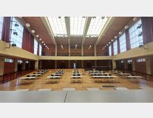 Großer Versammlungssaal mit einem Parkettboden, mehreren Reihen aus Tischen und Stühlen, hohen Decken und einer verglasten Lichtdecke. Links und rechts befinden sich große Fenster mit roten Vorhängen, und an der Stirnseite ist eine Empore erkennbar.