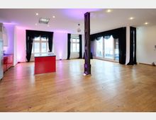 Ein heller, moderner Raum mit Holzboden, weißen Wänden und großen Fenstern, die von schwarzen Vorhängen eingerahmt sind. In der Mitte befindet sich eine rote Kücheninsel, und ein metallischer Stützpfeiler teilt den Raum optisch.