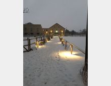 Ein schneebedeckter Weg führt zu einem großen Gebäude aus hellem Stein mit Satteldach, flankiert von Holzgeländern mit warmen Lichtern. Im Hintergrund sind schneebedeckte Flächen und Bäume erkennbar.