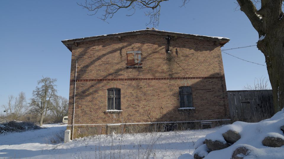 Zweistöckiges Backsteingebäude mit flachem Satteldach und vergitterten Fenstern in einer verschneiten Landschaft. Im Vordergrund liegen Schnee und Steine, daneben ein großer Baum; rechts schließt ein Holzzaun an.