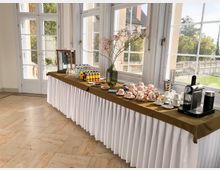Heller Veranstaltungsraum mit großen, weiß gerahmten Fenstern und Türen, die auf eine Terrasse und einen Parkblick führen. Entlang der Fenster steht ein langer Buffet-Tisch mit Tischdecke, Getränkeflaschen, Porzellan-Tassen und einer Kaffeemaschine.