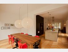 Moderne Wohnküche mit offenem Essbereich. Ein großer Holztisch mit roten und schwarzen Stühlen steht unter zwei runden weißen Hängeleuchten, daneben eine Kücheninsel aus Beton und dunklen Einbauschränken; der Raum hat helle Wände, geöltes Fischgrätparkett und große Fenster, die Tageslicht hereinlassen.