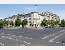 Straßenecke in einer Stadt mit mehrstöckigen, hellen Wohn- und Geschäftsgebäuden; im Vordergrund eine breite Kreuzung mit Markierungen, Laternen und Verkehrsschildern; im Hintergrund Bäume und ein U-Bahn-Zeichen.