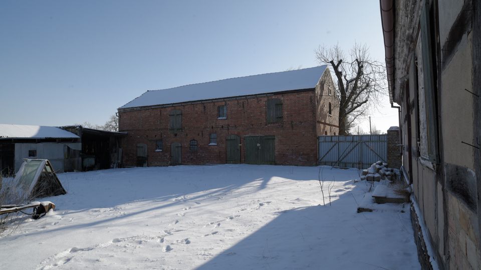 Verschneiter Innenhof eines Bauernhofs mit einer großen roten Backsteinscheune mit grünen Holztoren und wenigen kleinen Fenstern. Rechts begrenzt ein verwittertes Wirtschaftsgebäude den Hof, im Hintergrund stehen ein Metalltor und ein kahler Baum vor klarem Himmel.