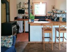 Kleine Wohnküche mit einem Holzofen links, einer hölzernen Theke mit zwei Barhockern in der Mitte und einem Fenster im Hintergrund. Die Küche ist mit offenen Regalen, Geschirr, und Küchenutensilien ausgestattet, auf der Theke steht ein Topf mit einer Pflanze.