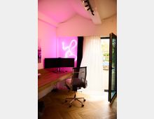 Ein modernes Arbeitszimmer mit einem Holzschreibtisch, einem ergonomischen Bürostuhl und zwei Monitoren. Im Hintergrund ist eine offene Balkontür mit weißem Vorhang und ein pink leuchtendes Neonlicht an der Wand zu sehen.