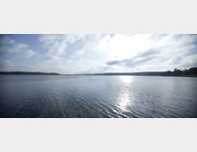 Weitläufige Wasserfläche eines Sees unter blauem Himmel mit vereinzelten Wolken, die Sonne spiegelt sich im Wasser; im Hintergrund sind bewaldete Ufer zu sehen.